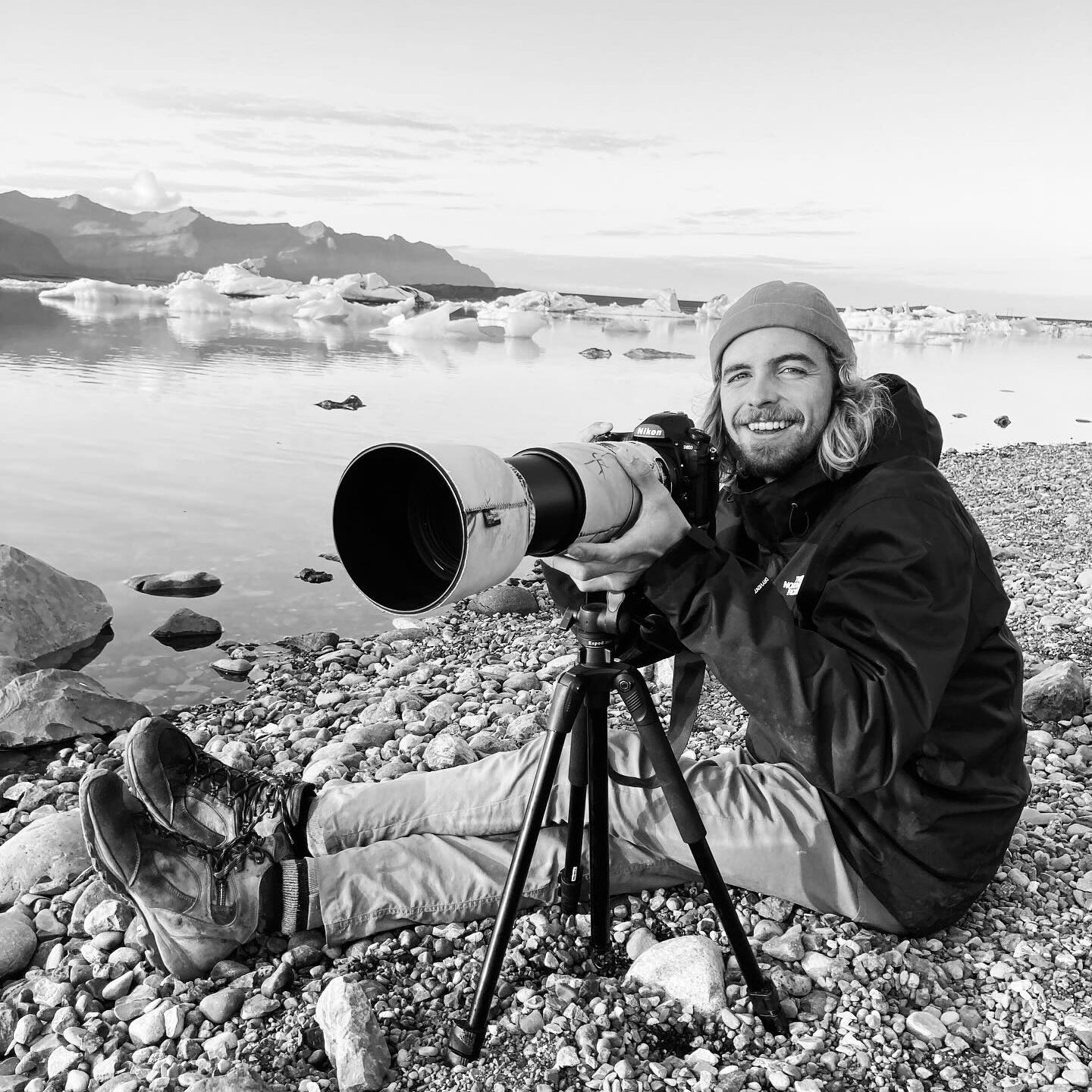 Photographe assis sur des galets au bord de l'eau, tenant un appareil photo équipé d'un objectif long, avec des montagnes et des icebergs en arrière-plan.